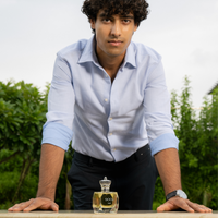 Male model leaning on a table with Tsar 900Y 100 ml perfume bottle placed in front, shot outdoors with greenery in the background
